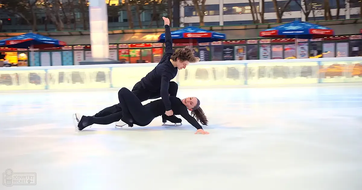 Young adult male and female figure skaters performing at an outdoor ice rink with trees and food stalls in the background, illuminated by evening light.
