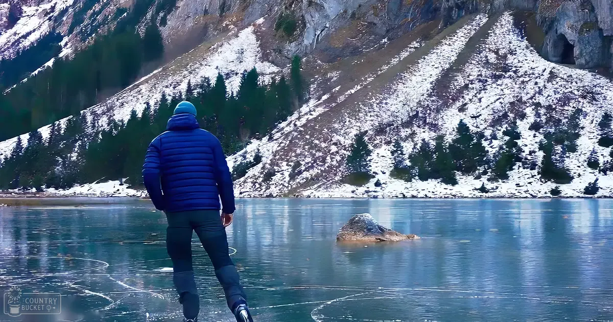 A male adult in a blue puffer jacket stands near a frozen lake surrounded by snow-covered mountains during winter.