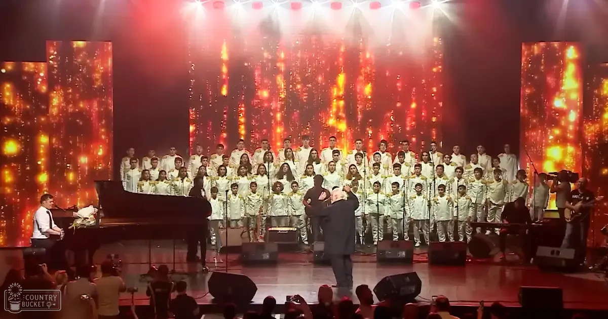 A choir performance with children and adults in white uniforms, a conductor in a dark suit, and musicians playing guitars under vibrant stage lights at an indoor concert hall.