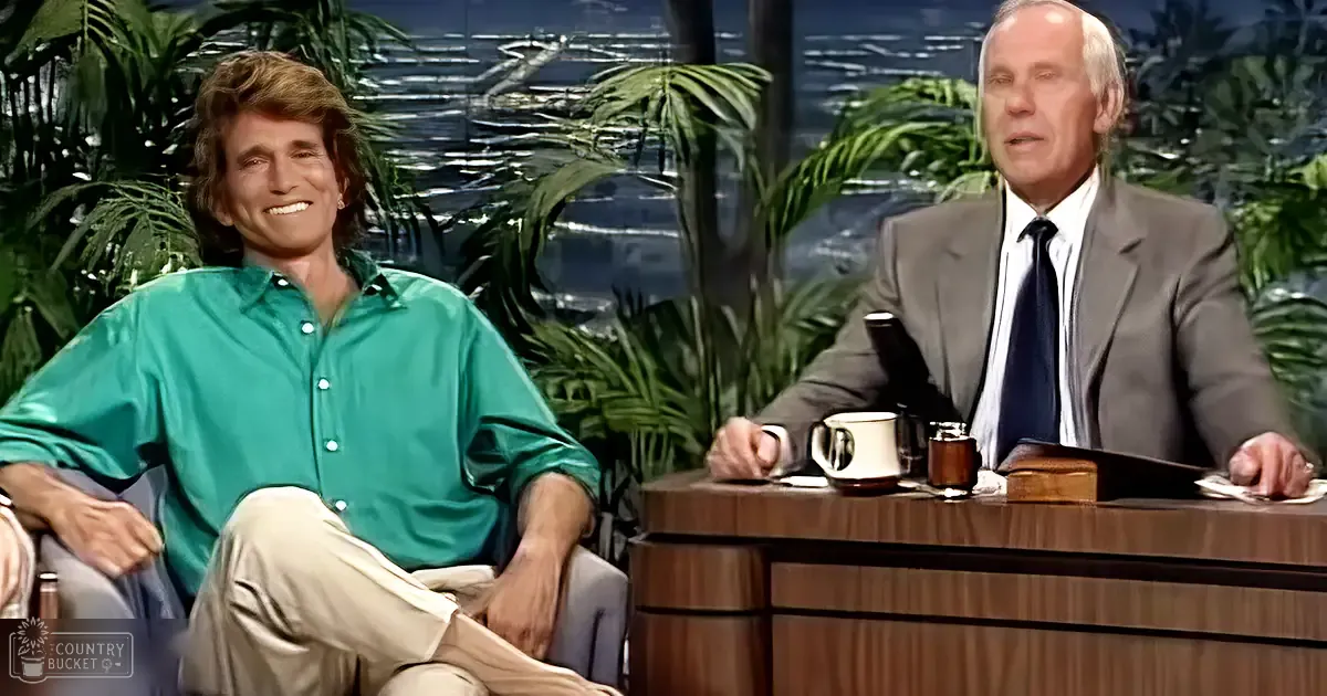 Two adult males on a talk show set in a professional studio with lush tropical plants, wearing casual yet stylish attire.