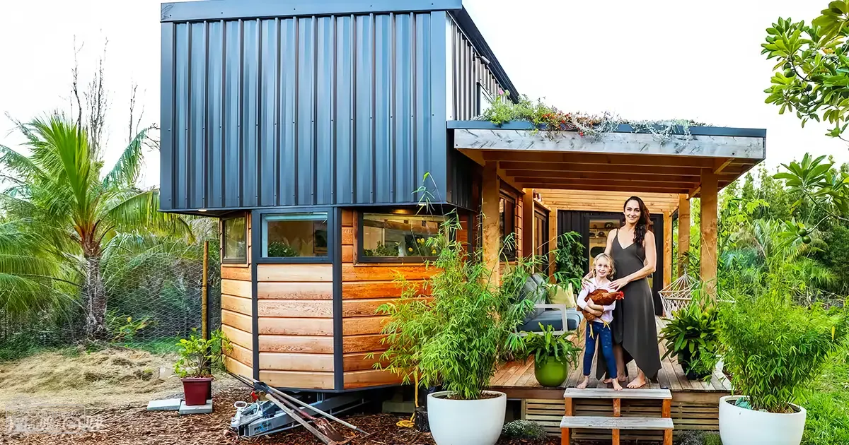 A woman with a toddler in front of a modern tiny house surrounded by greenery and potted plants under soft evening light.