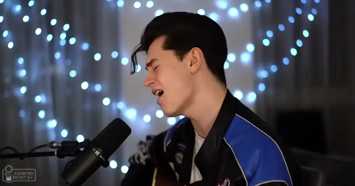 A young adult male in a relaxed fit black jacket with blue panels and white trim, holding a microphone indoors with string lights creating a bokeh effect behind him.