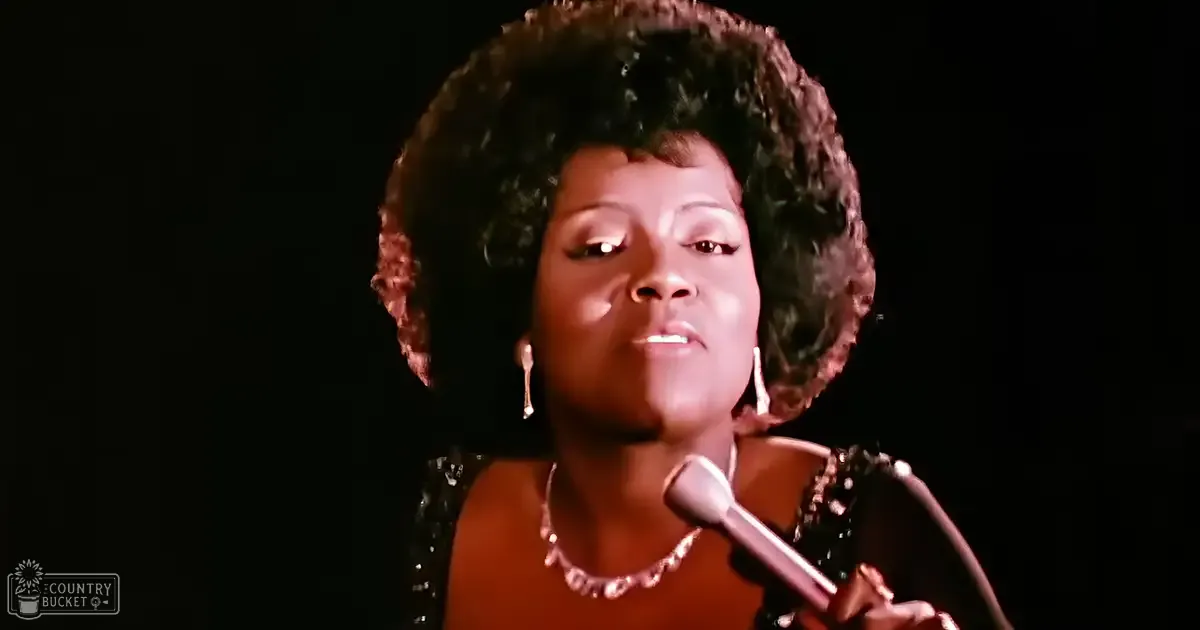 A female adult singer wearing an elegant black dress with sequins or beads, singing into a vintage-style microphone on a stage with controlled lighting and a dark background.