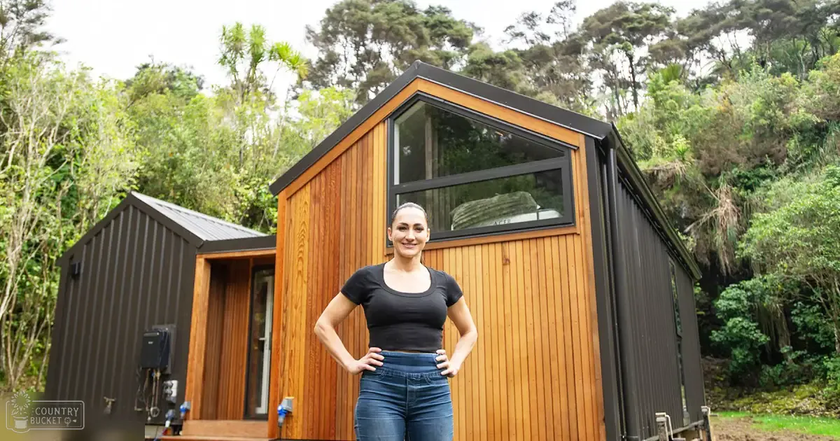 Adult female stands in front of a modern wooden cabin surrounded by trees, with overcast daylight illuminating the natural setting.