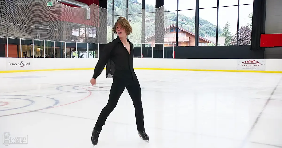 A young adult male figure skating in an indoor ice rink at Portes du Soleil, surrounded by a mountainous landscape with snow-covered peaks.