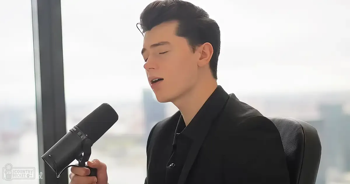 Young adult male in a black suit with a microphone indoors near large windows.