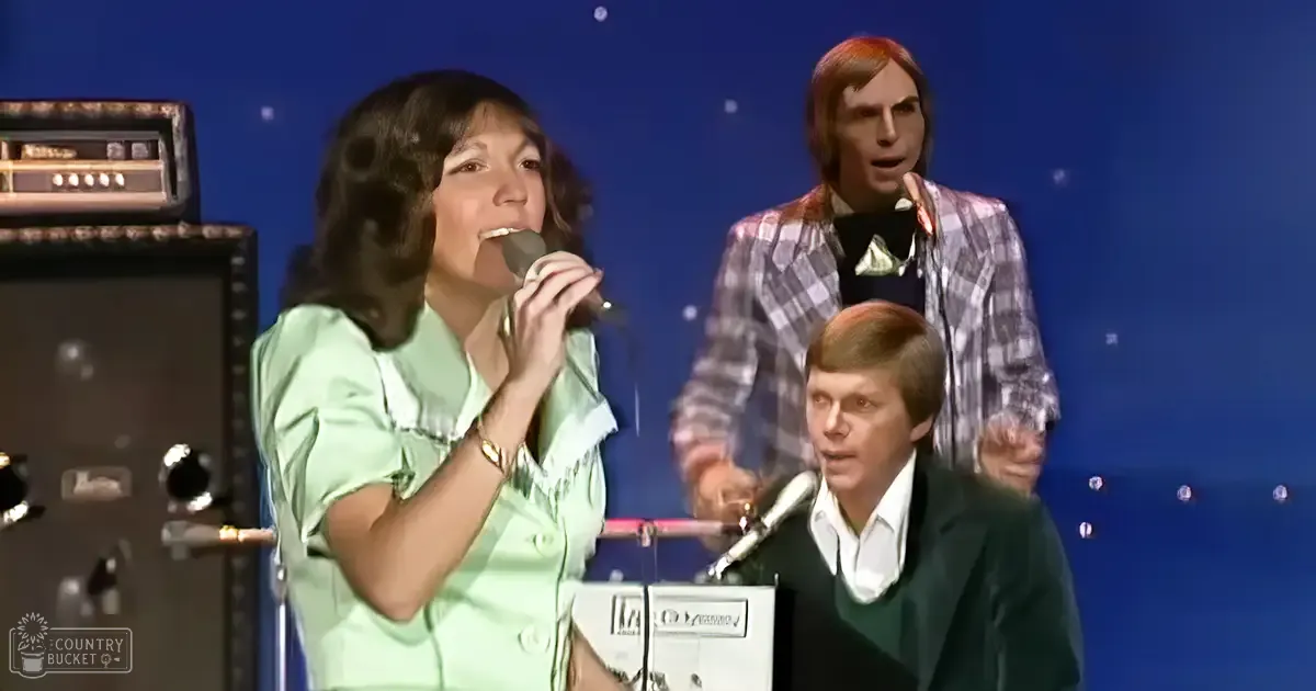 A woman singing into a microphone, wearing a light green ruffled blouse with a vintage 1970s aesthetic. A male in a plaid jacket and another in a dark sweater are present on stage.