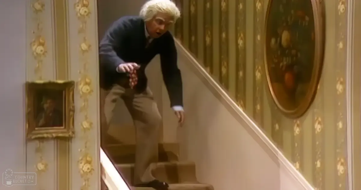 A middle-aged adult male in a tailored dark blazer, white shirt, light trousers, and black dress shoes stands indoors amidst ornate wallpaper with floral patterns and framed artwork. Daylight illuminates the elegant interior of what appears to be a grand home or mansion.