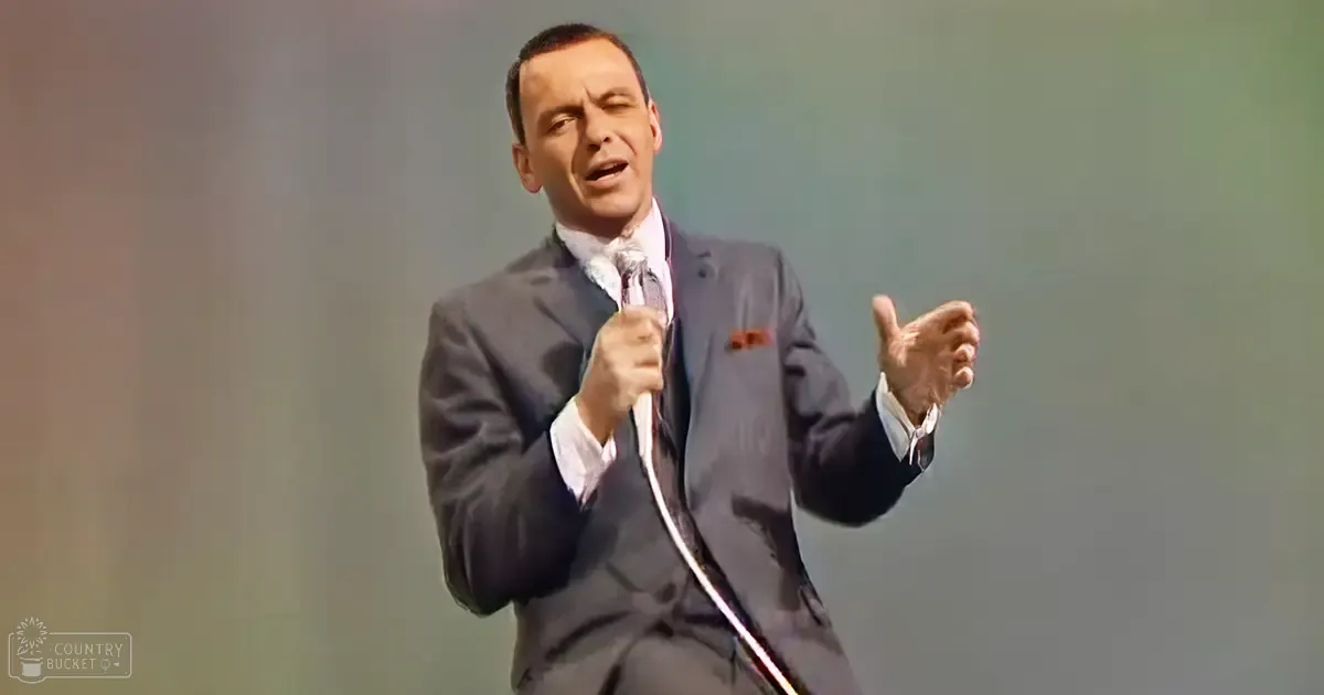 A middle-aged male singer in a dark gray suit, white dress shirt with an undone top button revealing part of his tie and light-colored tie, holding a microphone against a plain light-colored backdrop. The setting appears to be an indoor stage or performance venue.