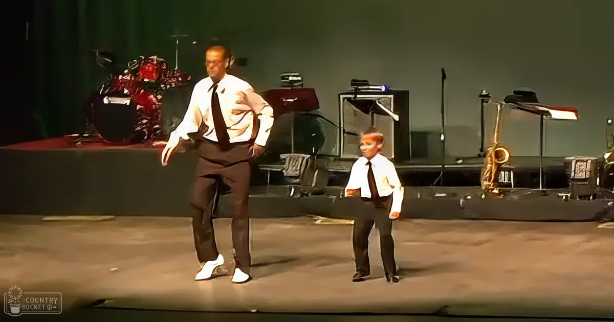 A middle-aged adult male in a white dress shirt and dark brown trousers performs on an indoor stage with a young child wearing similar attire, surrounded by musical instruments under stage lighting.