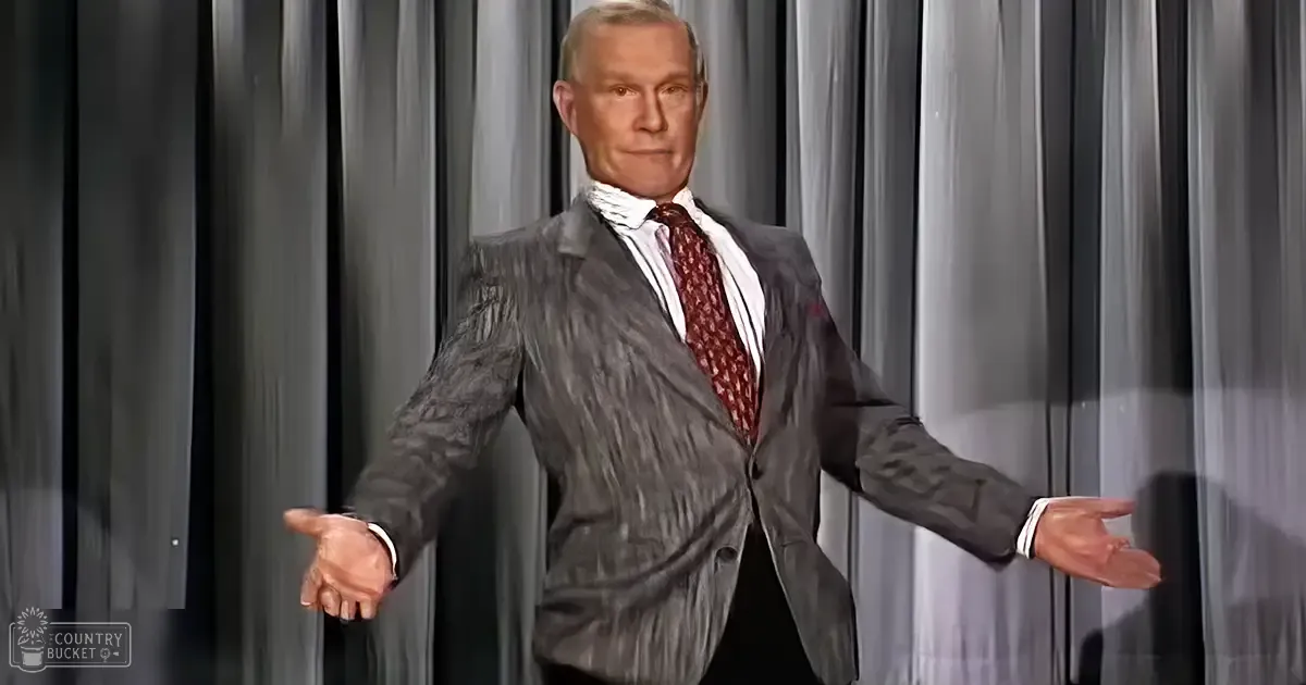 An older adult male in a gray pinstriped suit jacket and red patterned tie stands against a plain gray curtain backdrop, likely at an indoor performance venue.