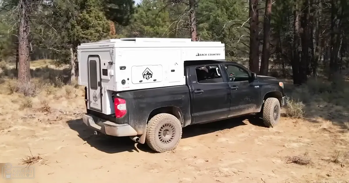 Black pickup truck with white overland camper on dirt path in forested area at dusk