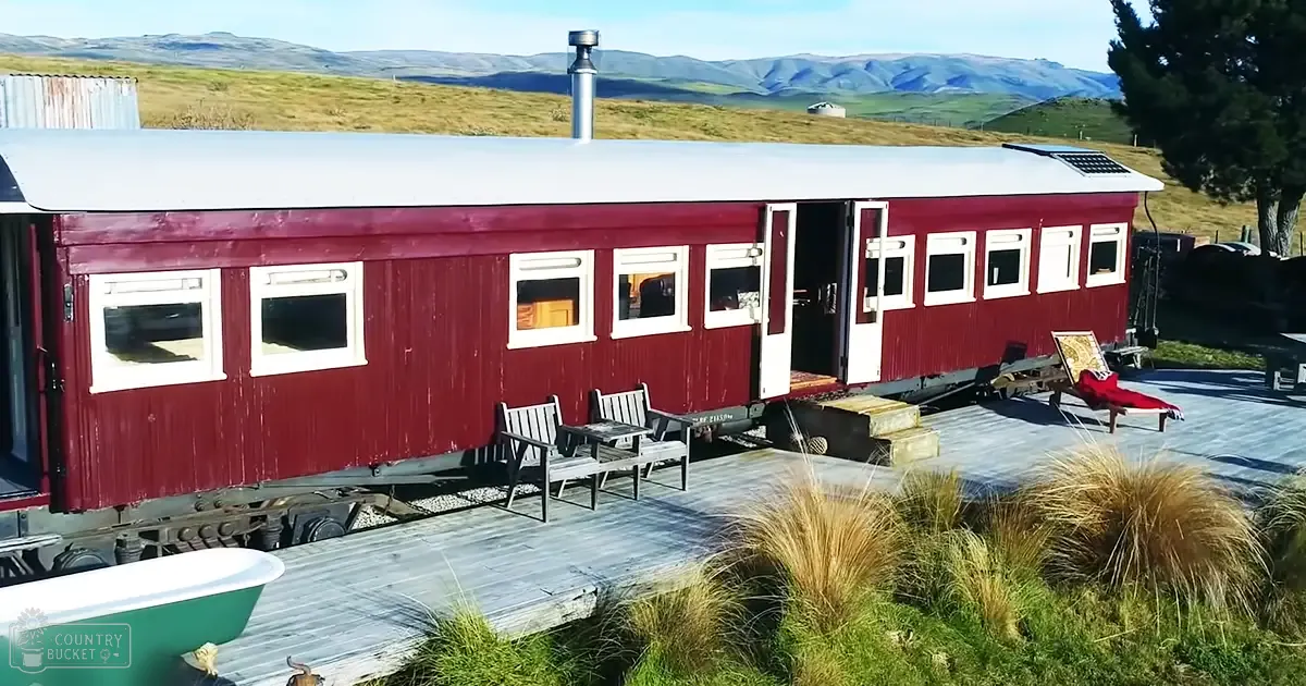 Rural setting with a repurposed train car converted into a mobile home, surrounded by rolling hills and mountains under sunny midday light.