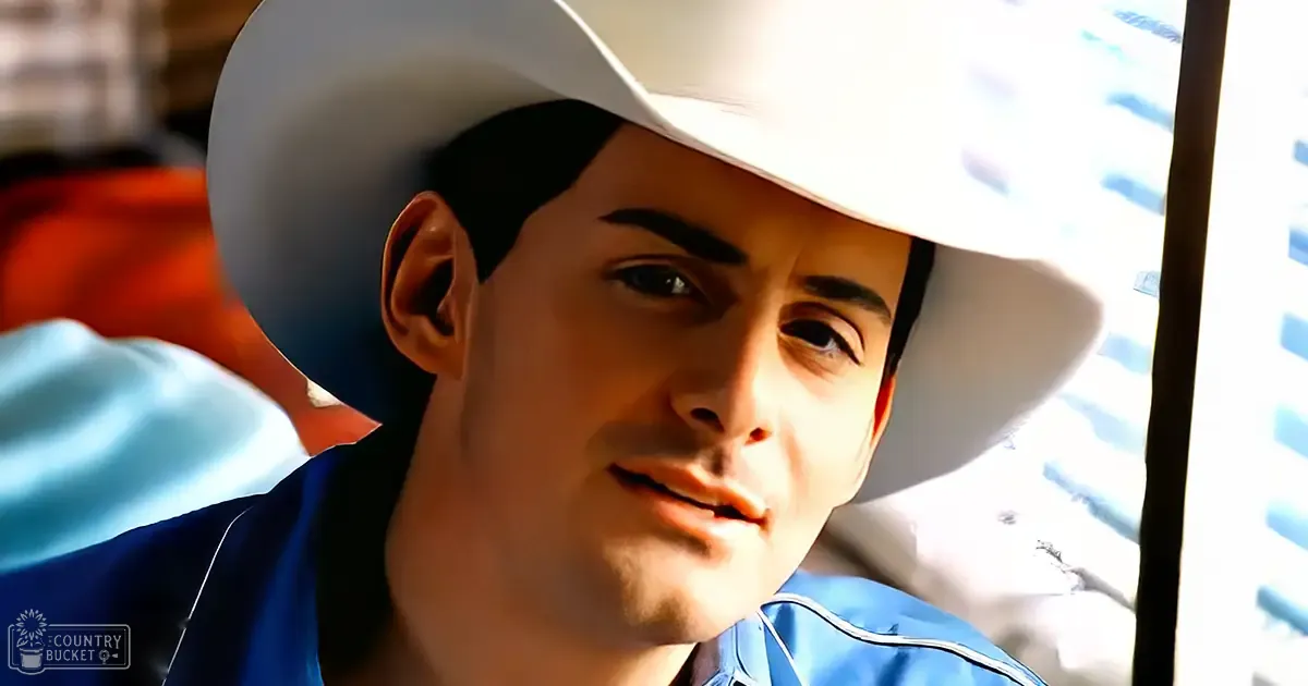 A young adult male wearing a bright blue shirt with white piping and a white cowboy hat stands indoors at what seems to be a social gathering, with blurred figures in the background.