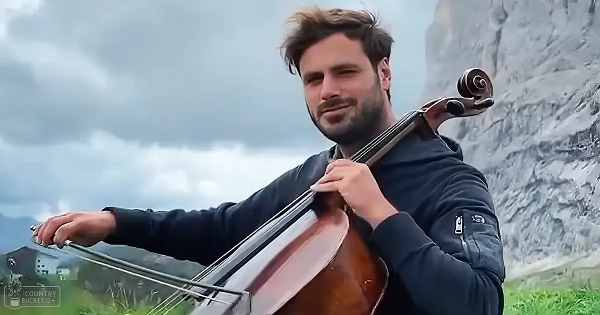 Adult male playing a cello in an outdoor setting with mountains in the background, suggesting late spring or early fall under diffused overcast light.