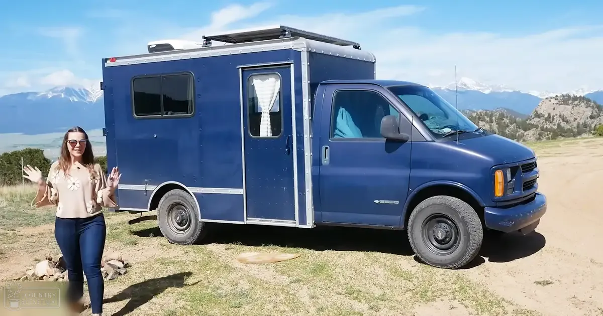 A young adult female wearing a beige floral blouse with bell sleeves, dark blue leggings or pants, and brown sandals stands in front of a blue Chevrolet camper van in an open field with snow-capped mountains under clear blue skies.