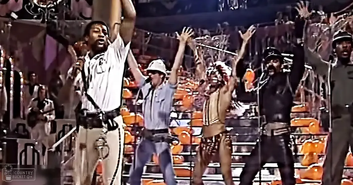 An adult male singer performs energetically with raised arms to an audience in a crowded indoor arena under artificial stage lighting, with rows of spectators and stage equipment visible.