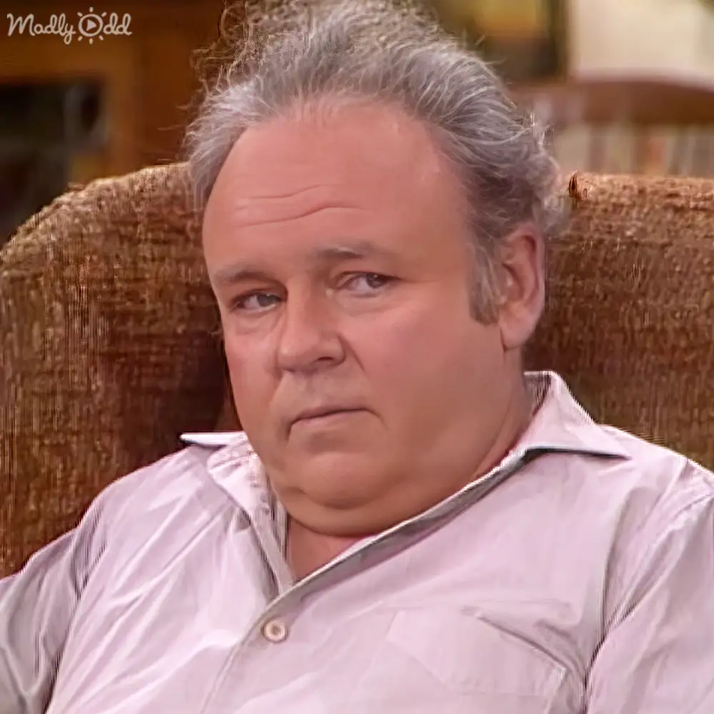 An older adult male seated in a brown upholstered chair, wearing a light pink button-up shirt with a collar and pocket on the left side of his chest, against an indoor background suggesting daytime lighting.