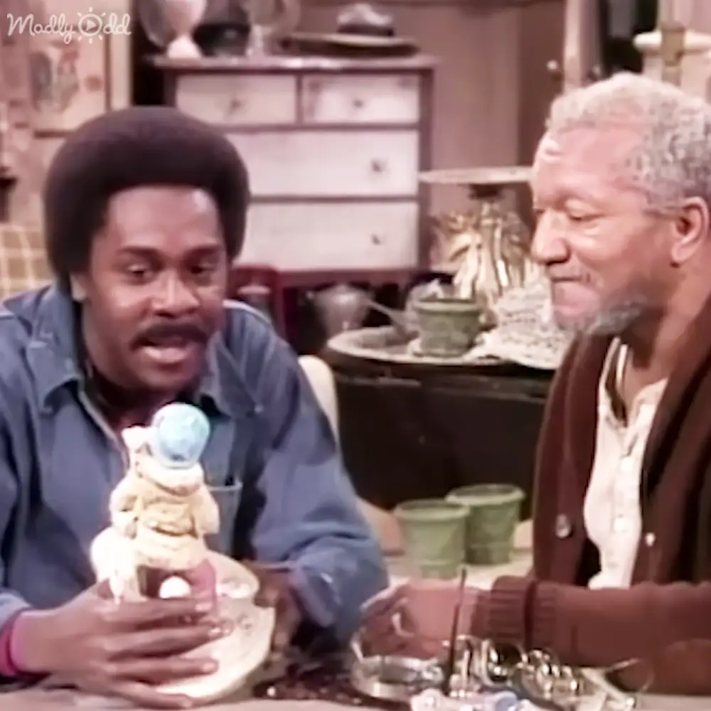 Two adults interacting casually indoors, suggesting a cozy living room scene with furniture and decorative items.