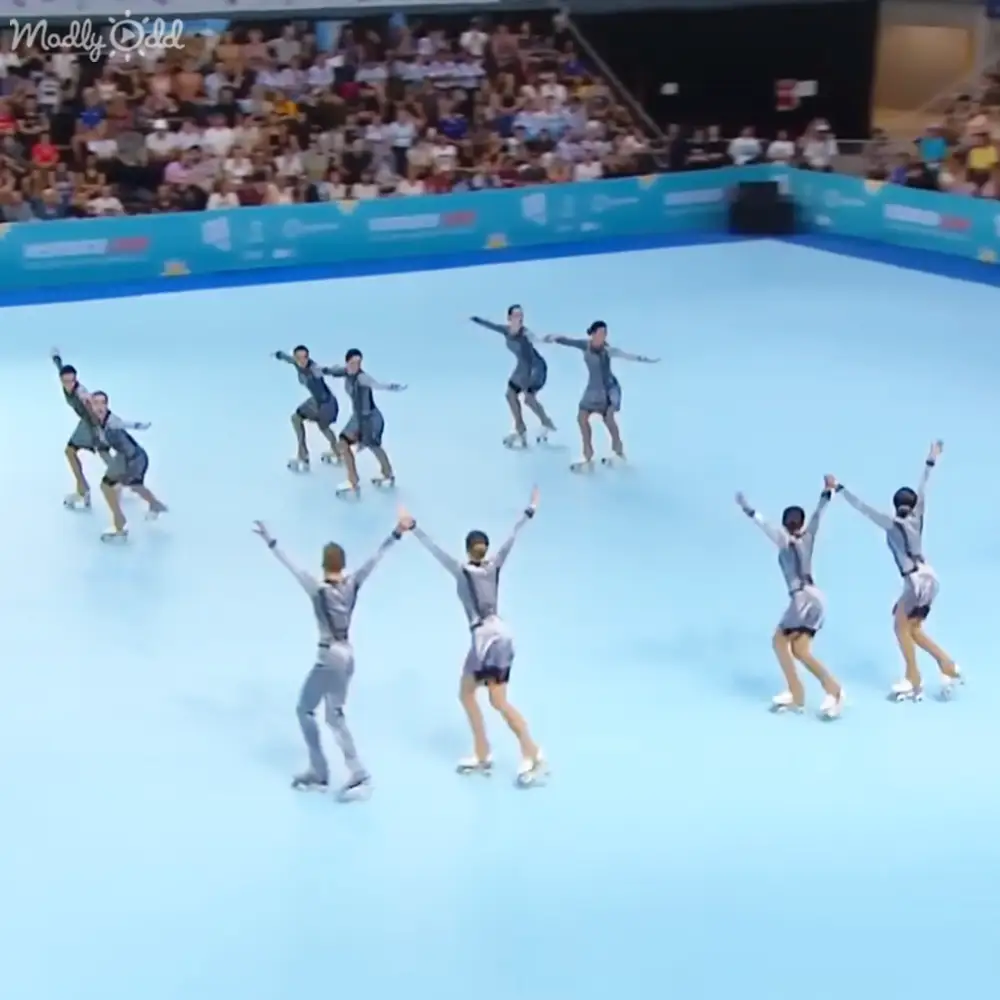 A synchronized skating team performing at an indoor ice rink with spectators watching from stands, set against blue walls with advertisements.