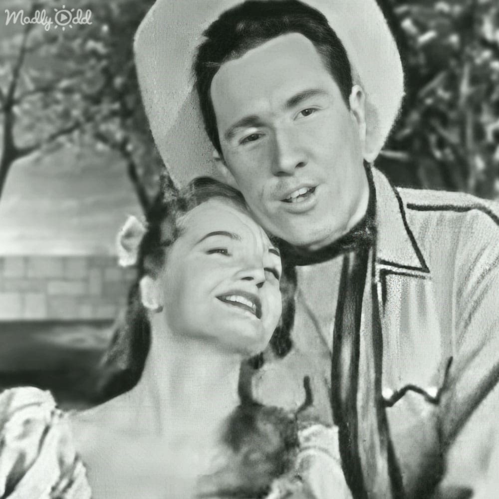 A couple in a Western setting, dressed in early-to-mid-20th-century attire, standing outdoors on what appears to be a porch or patio with greenery and soft natural light.