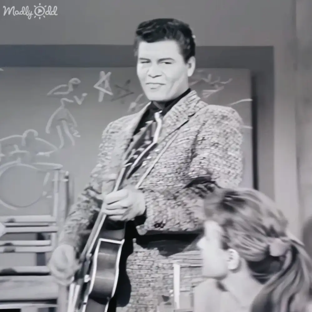 A young adult male performer holding a guitar in a studio setting with a child in the foreground.