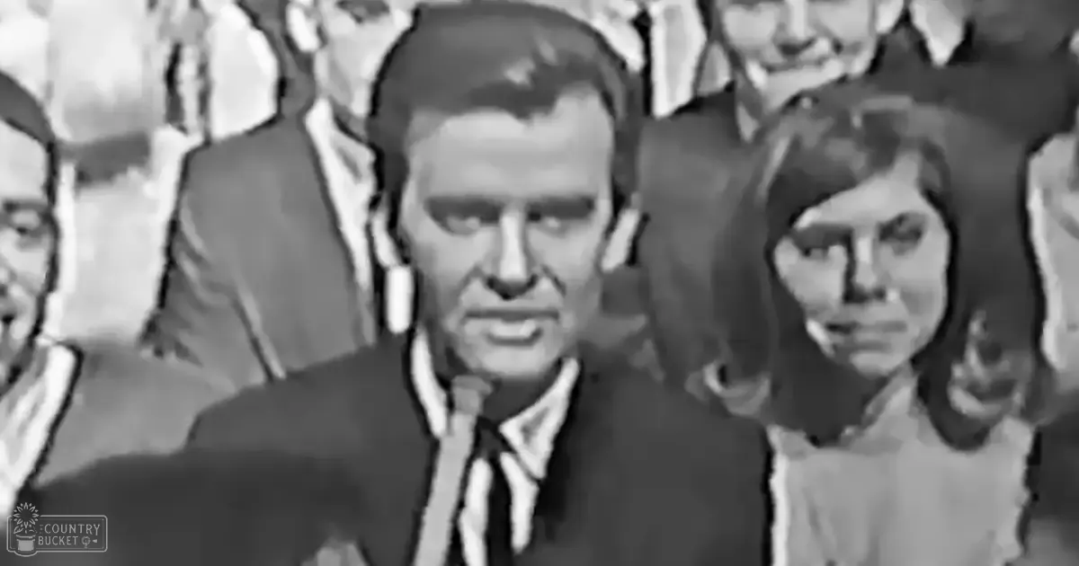 Adult male wearing a dark suit jacket and tie at an indoor event with other adults around.