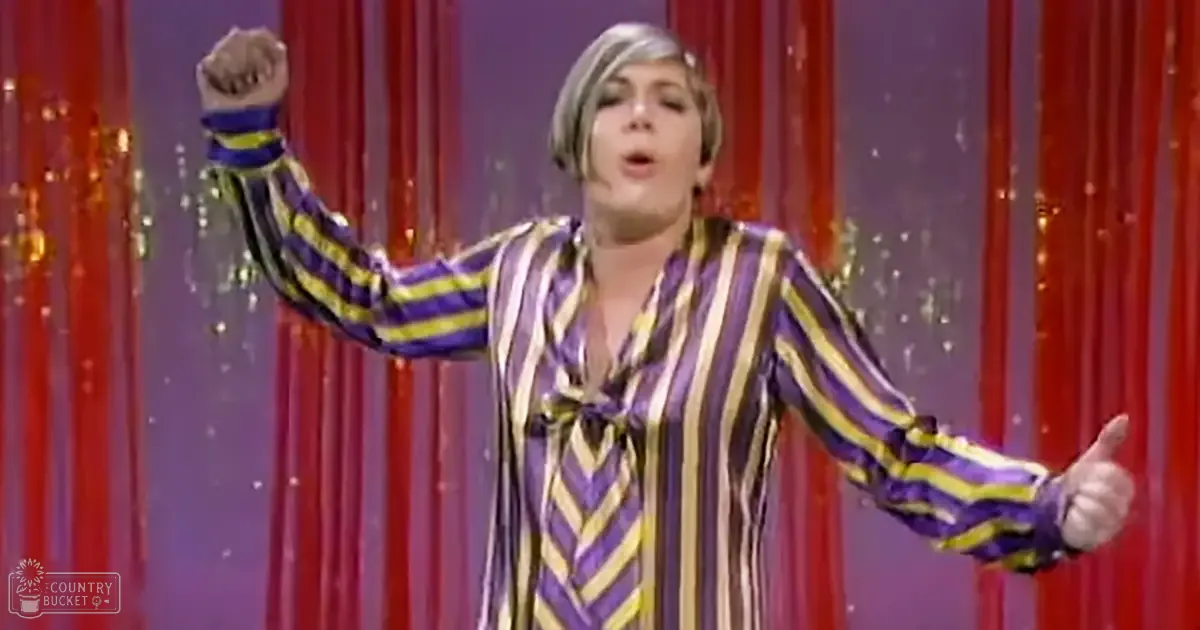 An adult female performer in a studio setting, wearing a loose-fitting, striped shirt with purple, yellow, white, and black vertical stripes. The backdrop includes red curtains with decorative gold tassels.