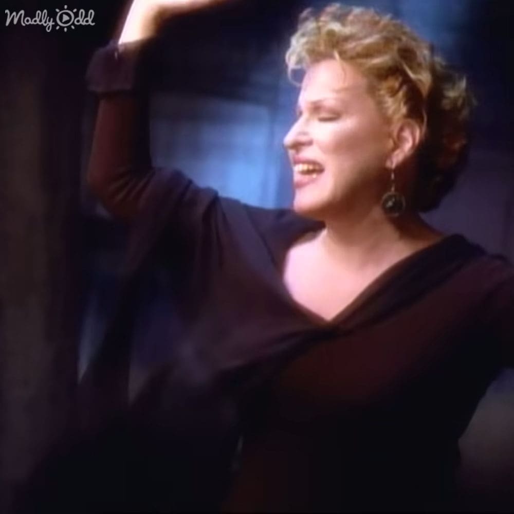 A woman wearing a black, asymmetrical deep V-neck dress with rolled-up sleeves and large hoop earrings stands indoors under dim lighting.