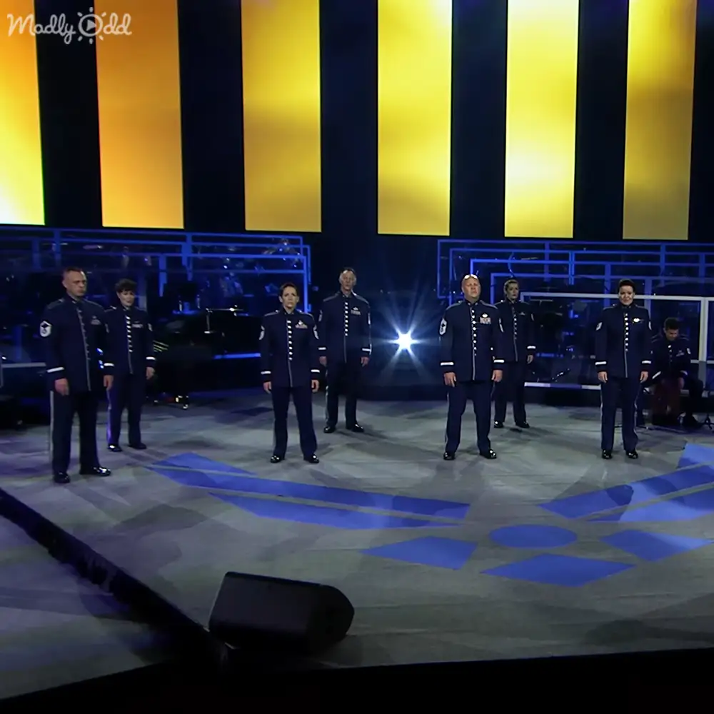 A group of uniformed military personnel performing on stage in a concert hall or theater with bright stage lighting.