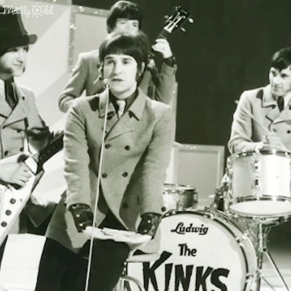 A young adult male singer in a music studio, wearing a double-breasted coat from the 1960s era, holding a microphone while singing into it. Other band members are also present, dressed similarly.