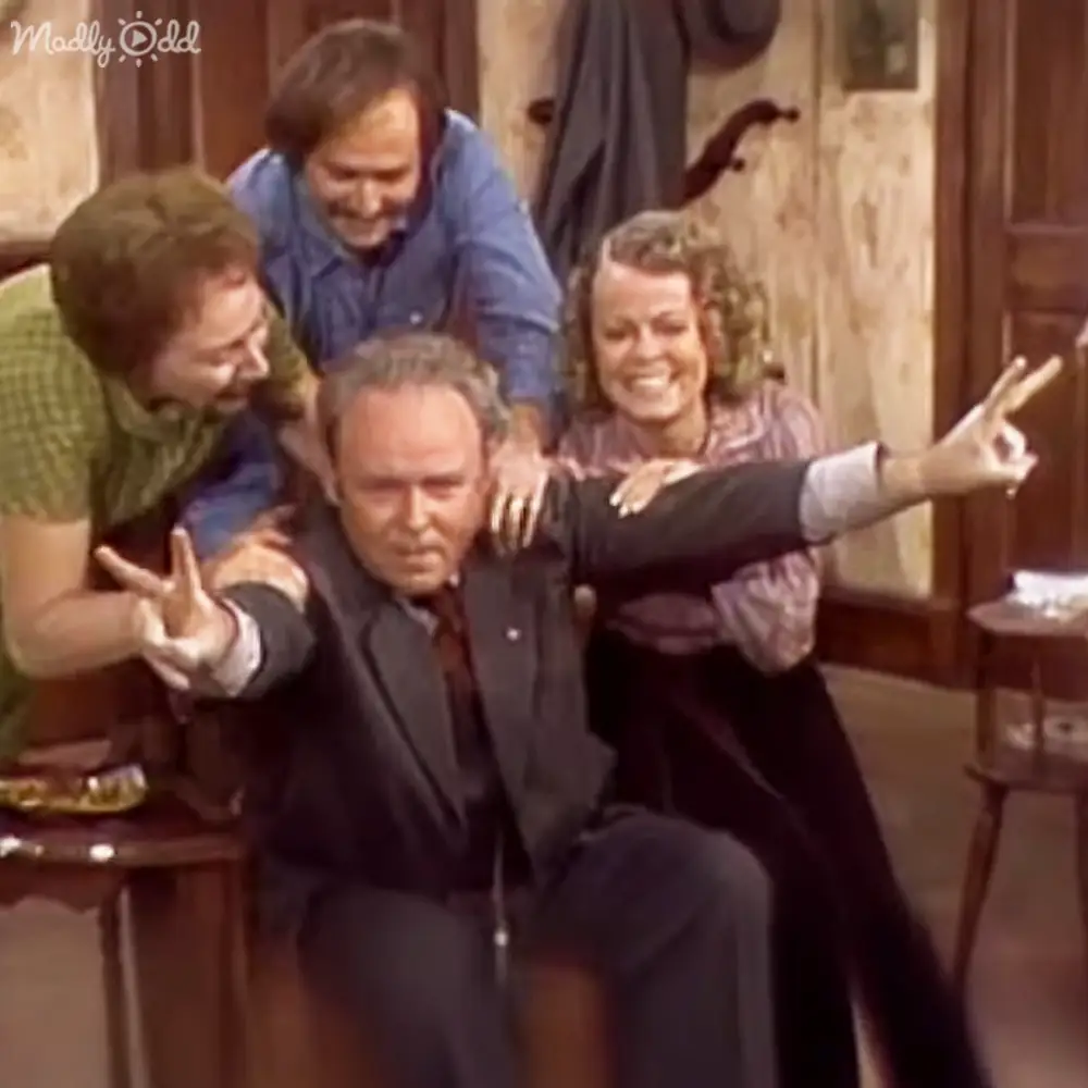 A middle-aged man in a dark suit seated at the center surrounded by three other individuals in casual attire within an indoor setting with rustic wooden furniture and warm lighting.