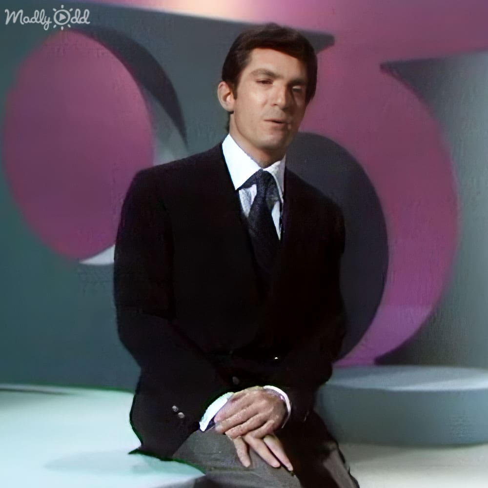 A young adult male in a formal black suit and collared shirt with tie, seated on a white platform indoors with abstract geometric shapes in the background.