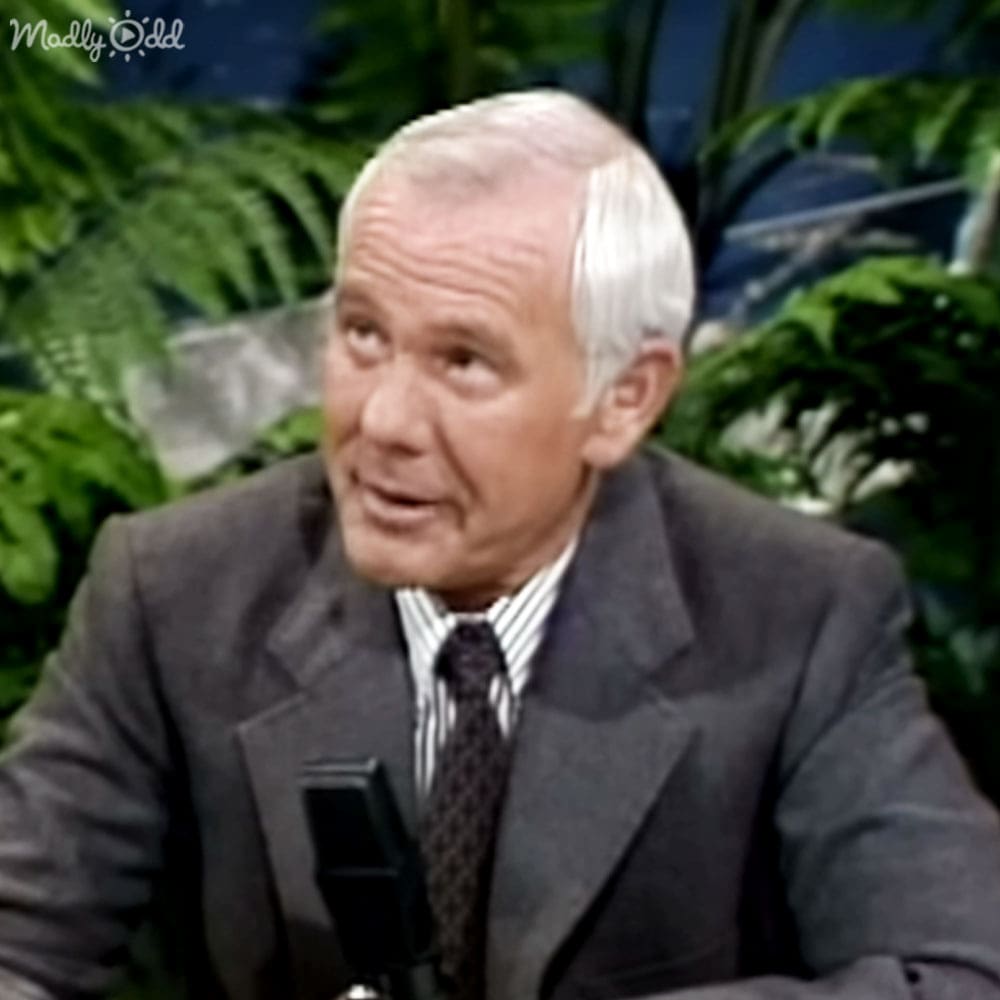 Older adult male in a studio setting with plants and microphone, dark gray suit jacket, white collared shirt, black tie, even bright lighting.