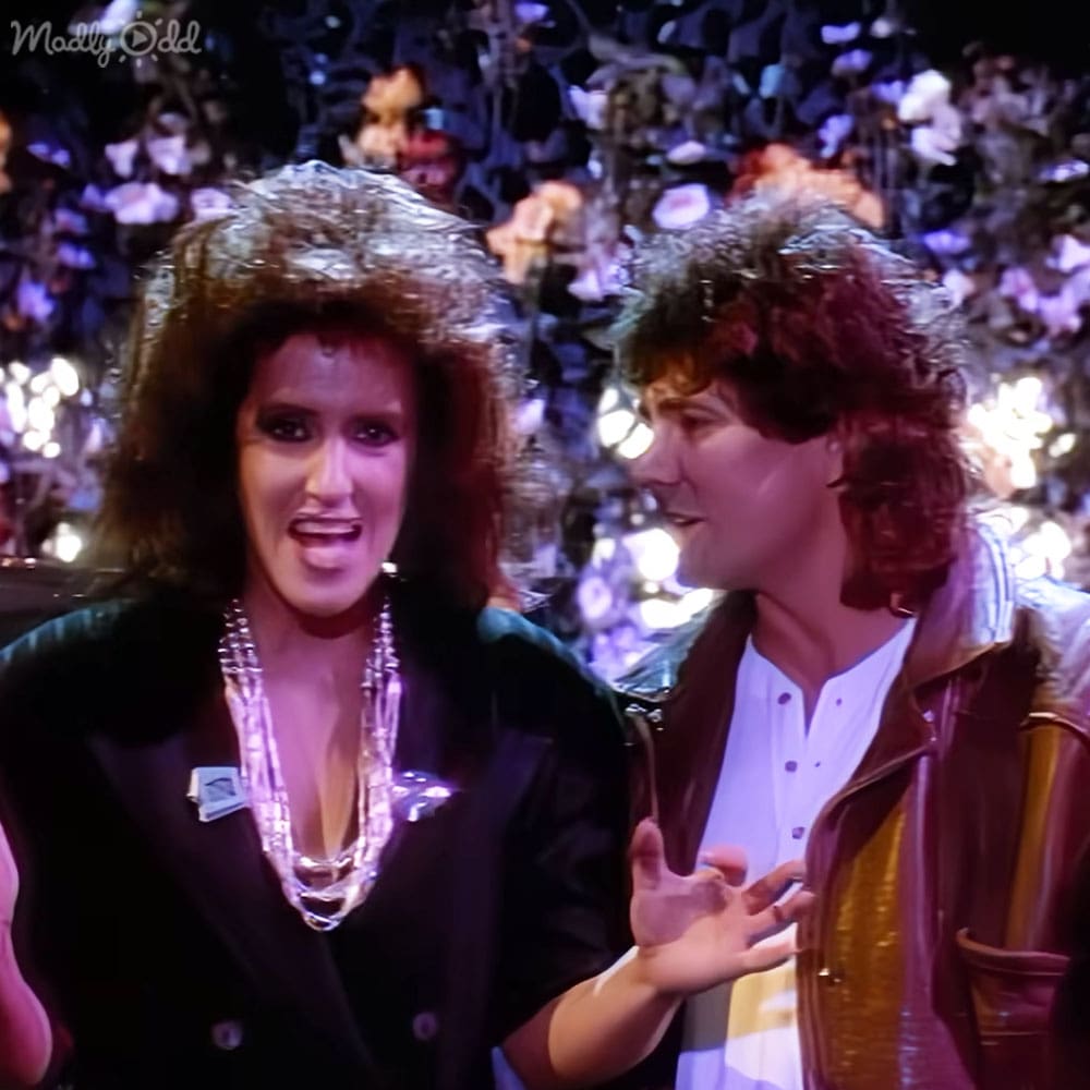An adult female in a black blazer with metallic embellishments at an event with a crowd; next to her, an adult male in a brown leather jacket and white shirt.