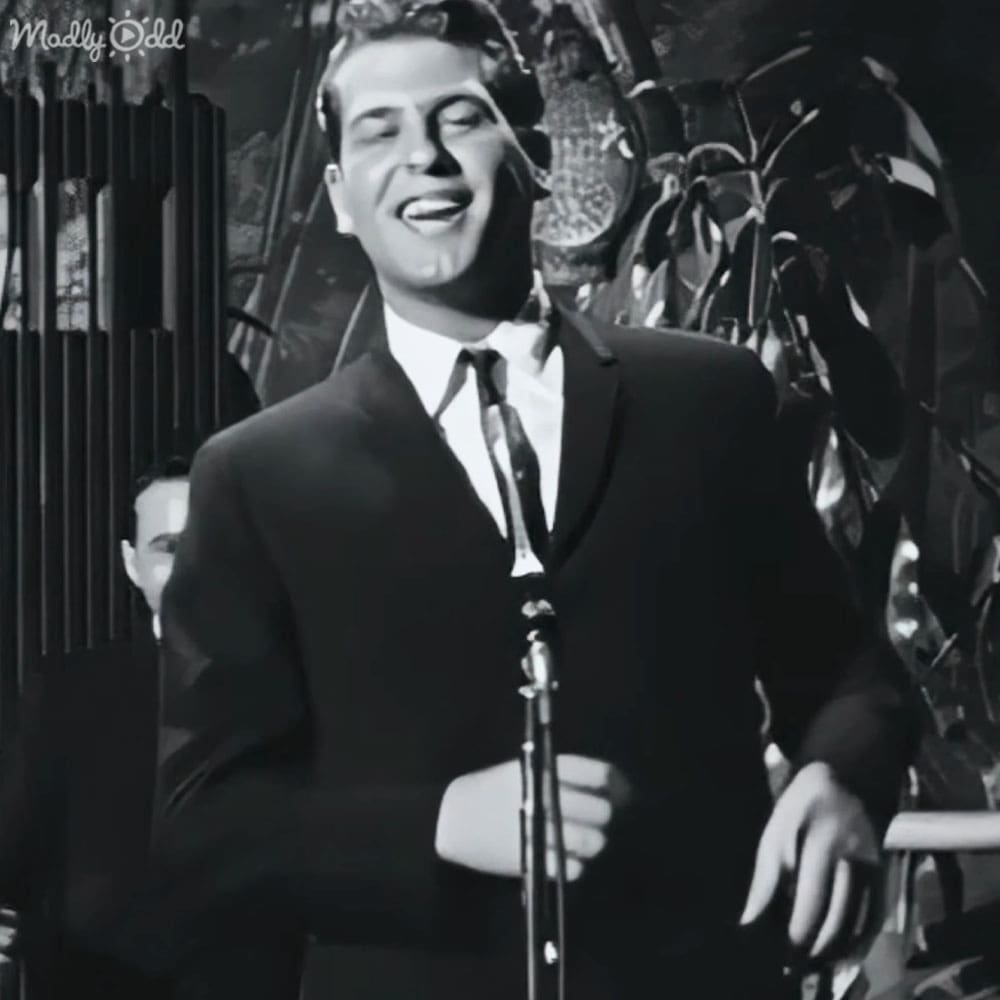 A young adult male singer wearing a dark suit with a white shirt and tie, performing on stage under focused lighting at night.