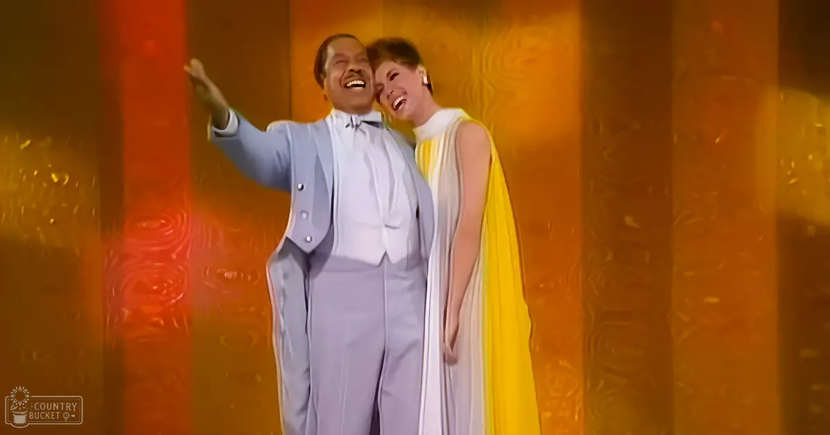 Adult male singer in a light blue suit with classic mid-20th-century style, performing alongside an adult female in a dramatic, striped sleeveless dress against a vibrant orange and yellow textured backdrop.