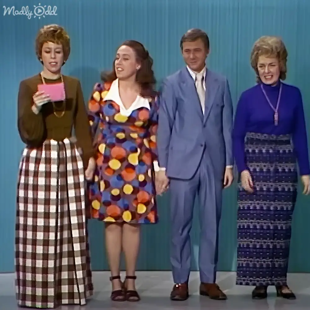 Four individuals in a mid-20th century event setting with plain blue backdrop, adult females and males wearing vintage clothing.
