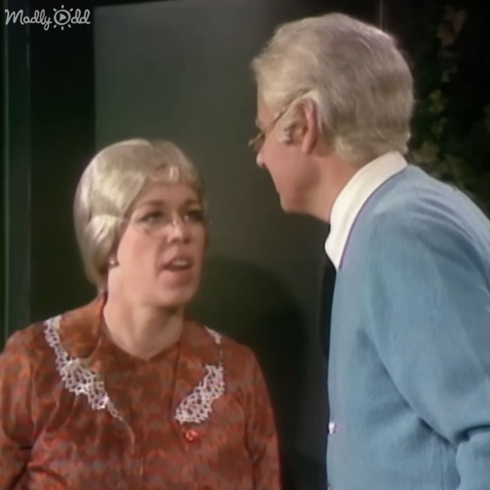 A middle-aged woman wearing a redish-brown blouse with lace detailing, standing indoors with an older adult male in a light blue cardigan. Neutral-colored walls and natural daylight suggest daytime in a simple domestic setting.