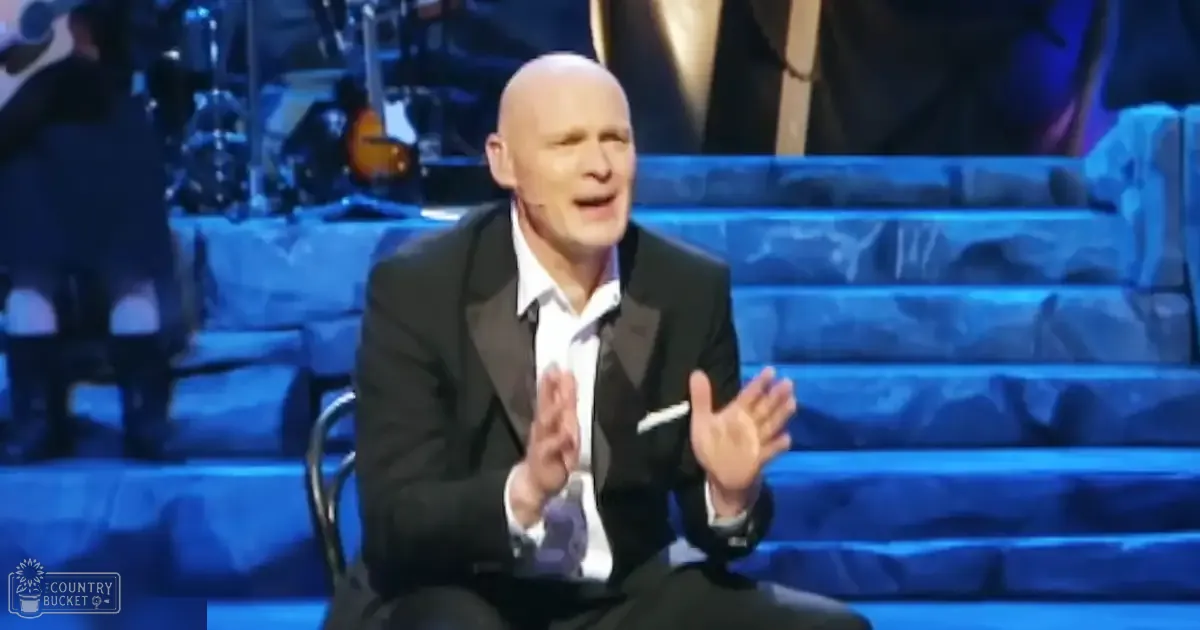 A middle-aged to older adult male wearing a black suit with a white shirt and pocket square, seated on a chair in front of blue steps under stage lighting at an indoor venue.