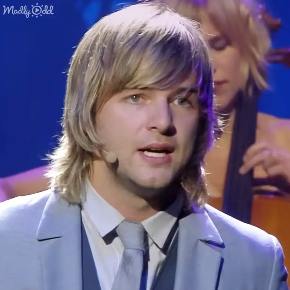 Young adult male in a light blue suit jacket and white shirt with dark tie stands on an indoor stage or performance area under artificial lighting, possibly evening or night.