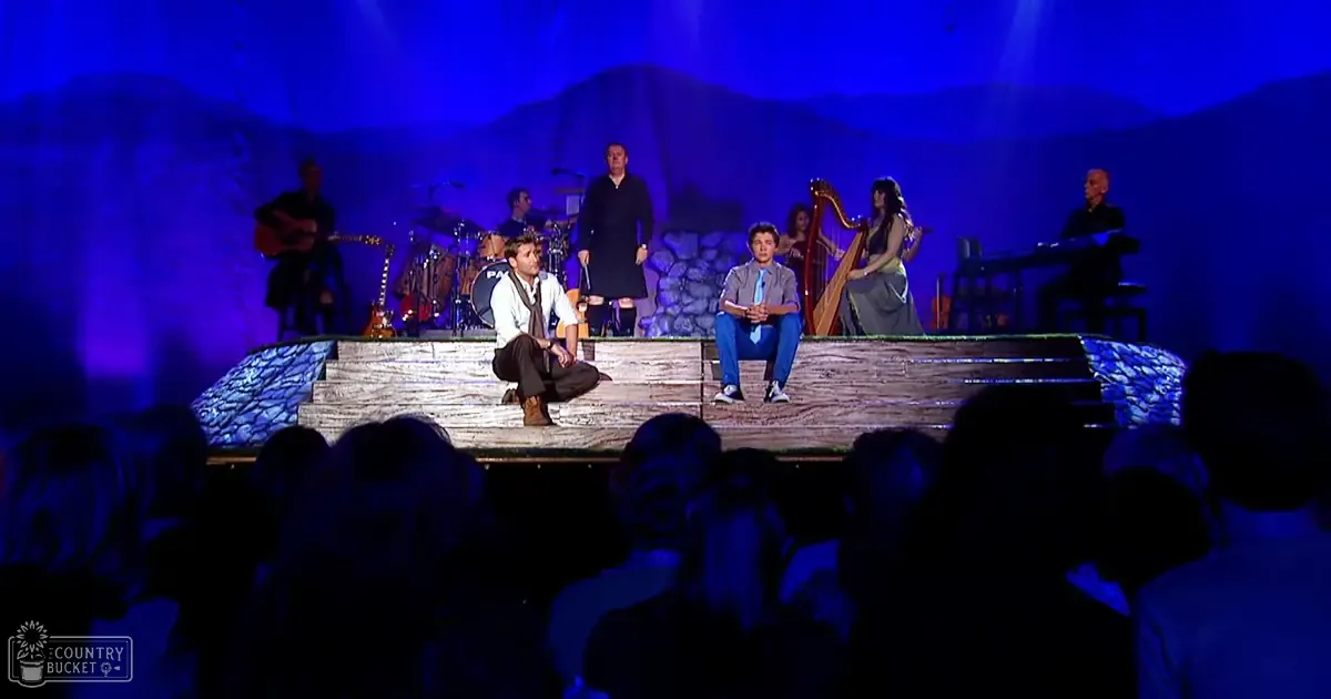 A live musical performance with a rustic, natural-themed stage setup under blue lighting against a backdrop suggesting a mountainous landscape.