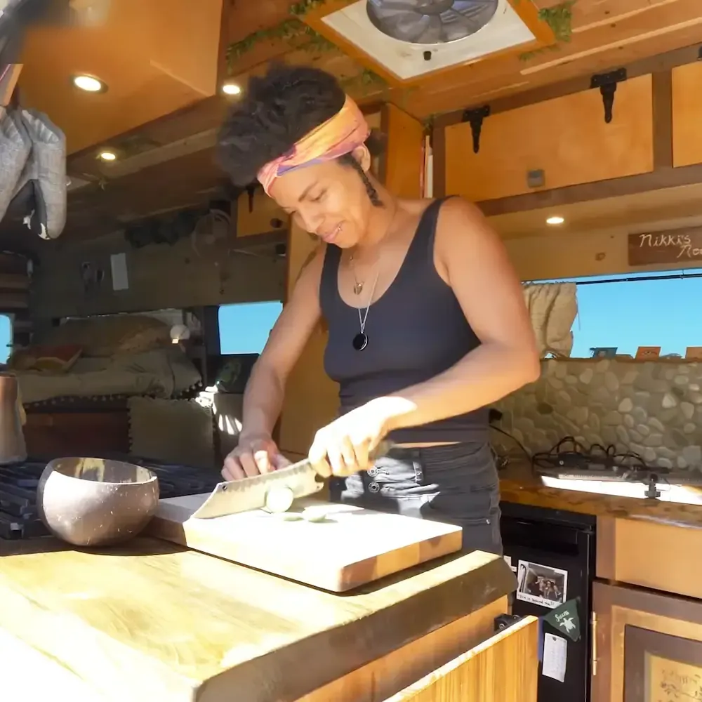 An adult female wearing a black scoop-neck tank top and dark jeans with colorful headband and pendant necklace inside an RV kitchen during daytime.