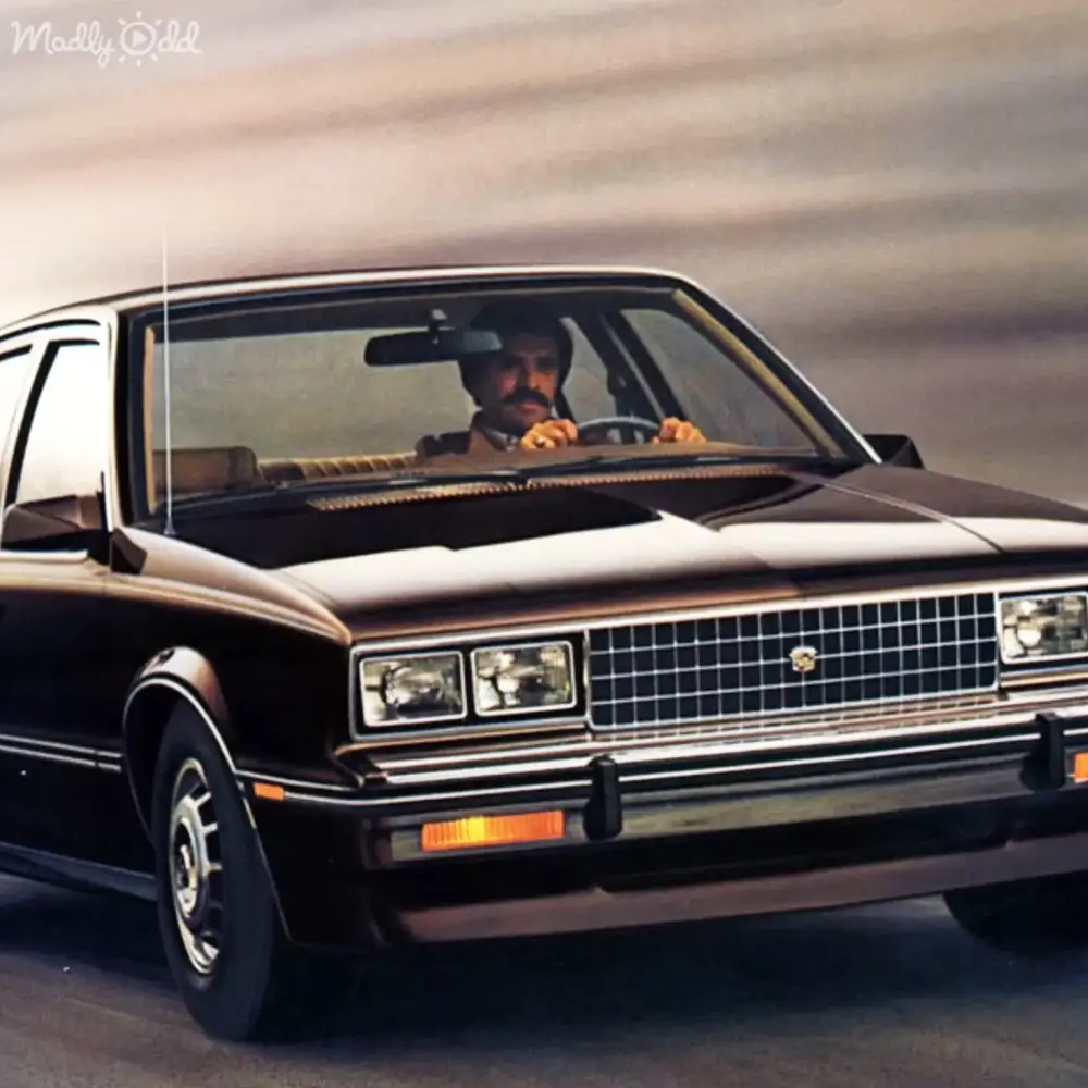 A middle-aged man in a dark-colored leather jacket drives a retro-styled black car on an open road under cloudy skies, suggesting late spring or summer.