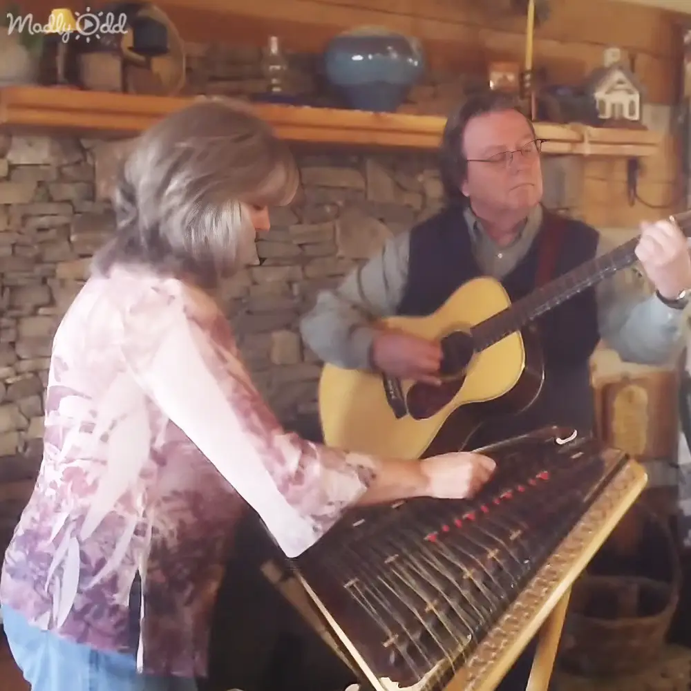 A musical performance in a cozy indoor setting with a stone fireplace, wooden shelves, decorative vases, small sculptures, and a dulcimer.