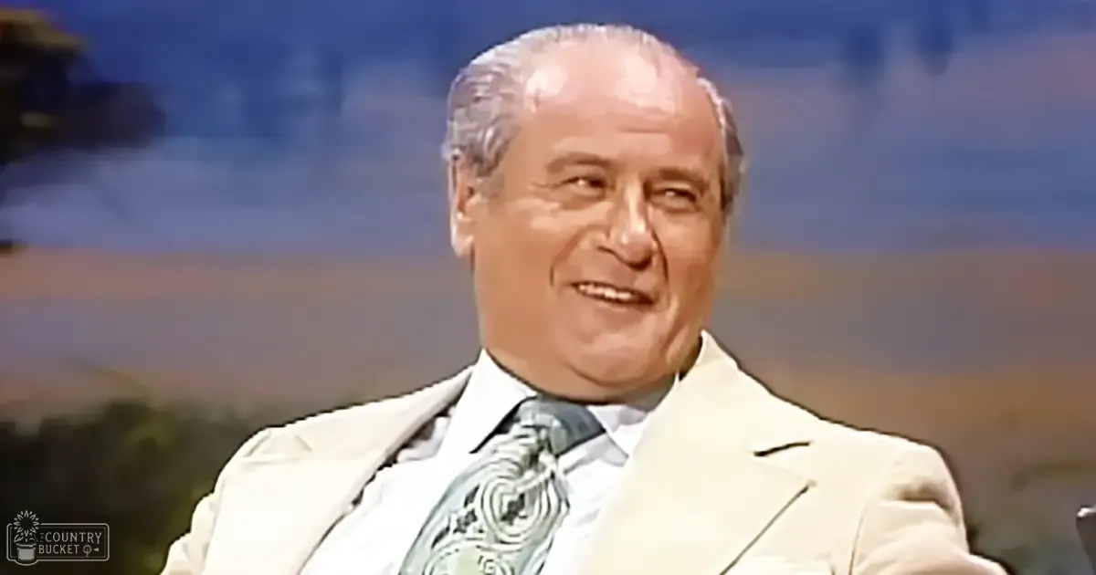 An older adult male wearing a light-colored suit jacket with a patterned tie stands against a blurred outdoor backdrop suggesting a studio setting.