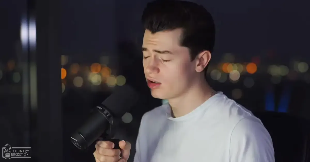 A young adult male singer wearing a white, relaxed fit crew-neck shirt stands in an urban nighttime setting with blurred city lights behind him.