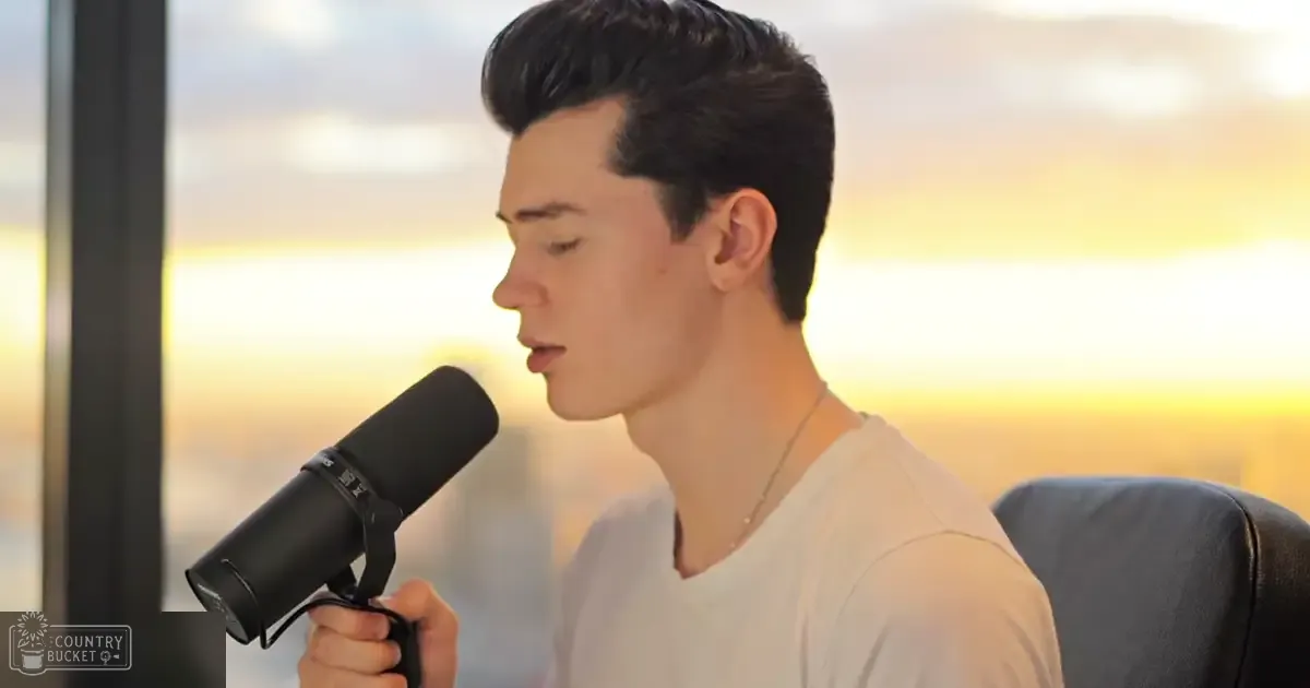 Young adult male wearing a white V-neck t-shirt, holding a black microphone in an indoor setting with a scenic sunset view outside.