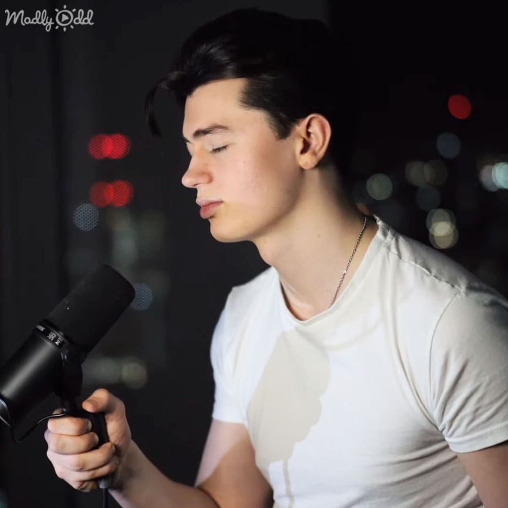A young adult male wearing a white t-shirt with a subtle design, holding a black hairdryer indoors.
