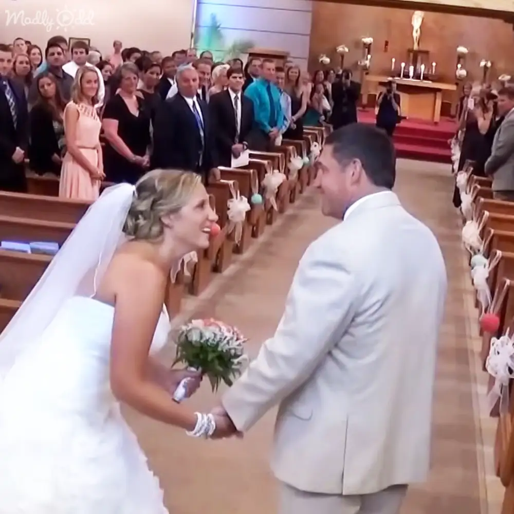 A bride in a white wedding dress with a veil and bouquet stands beside her groom wearing a light suit jacket indoors at an altar in a church setting.